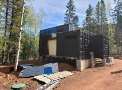 Modern black shipping container home in forest clearing with blue awning