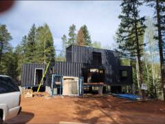 Black tiny house on wooden deck surrounded by evergreen trees and parked vehicles
